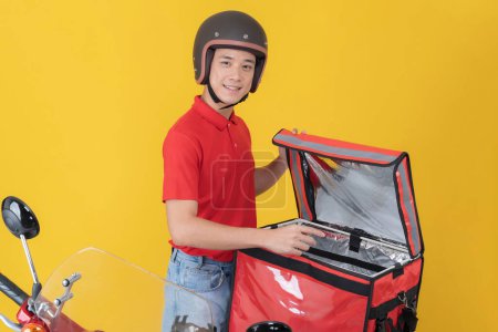 Smiling young male delivery man in a red shirt and helmet stands beside a motorcycle, opening an insulated delivery box against a vibrant yellow background, ready to dispatch an order