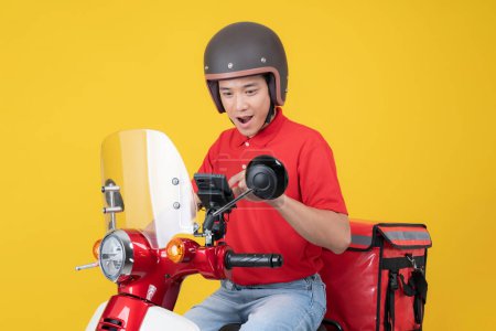 Young asian delivery man in a red uniform and helmet, checking his smartphone while sitting on a red and white motorcycle against a vibrant yellow background