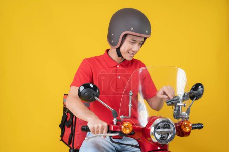 Young asian delivery man in a red uniform and helmet, checking his smartphone while sitting on a red and white motorcycle against a vibrant yellow background