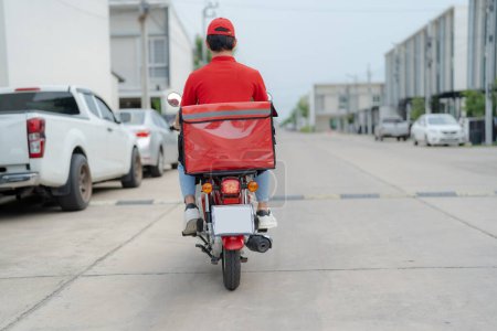 Foto de El repartidor con uniforme rojo y casco monta una moto por una calle urbana, con vehículos y edificios en el fondo, ejemplificando los servicios de entrega de la ciudad - Imagen libre de derechos