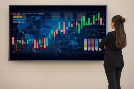 Professional businesswoman stands in front of a large digital screen displaying colorful stock market charts and data for financial analysis and strategic planning in a corporate environment
