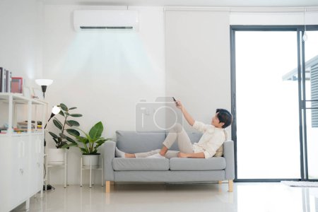 Le jeune homme est assis sur un canapé, contrôlant confortablement la climatisation avec une télécommande, dans un salon moderne et bien éclairé, présentant un style de vie de commodité et de technologie