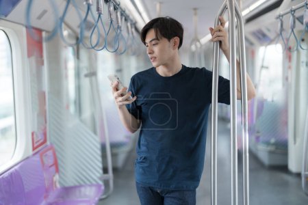 Jeune homme asiatique se tient dans un train de métro, tenant un poteau et se concentrant sur son écran de smartphone tout en se rendant au travail. L'environnement urbain moderne se reflète dans le chariot propre et vide
