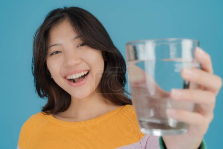 Foto de Mujer joven feliz con una sonrisa brillante sosteniendo un vaso de agua transparente con confianza, vestido con un suéter a rayas de colores, de pie sobre un fondo azul sólido - Imagen libre de derechos