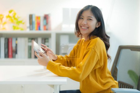 Foto de Joven mujer asiática alegre se involucra con su teléfono inteligente, cómodamente sentado en un elegante escritorio blanco en una oficina en casa brillante y bien organizada con estanterías en el fondo - Imagen libre de derechos