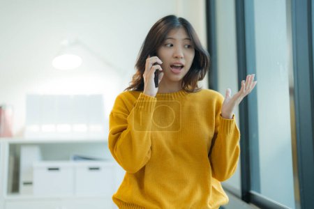 Mujer joven en un suéter amarillo que se ve sorprendida mientras tiene una conversación en su teléfono móvil, de pie en el interior con un fondo interior moderno
