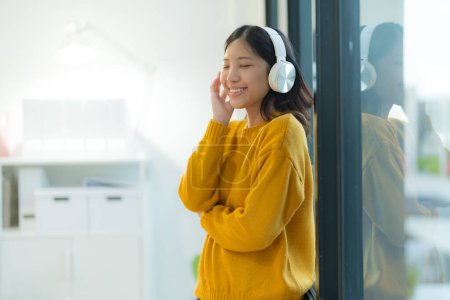 Comfortably dressed in a cozy yellow sweater, a young woman smiles as she enjoys her favorite tunes on headphones, standing by the window in a bright, modern home interior