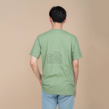 Stylish young man poses with confidence in a plain green t-shirt and blue jeans against a neutral beige background, perfect for fashion and apparel mockups