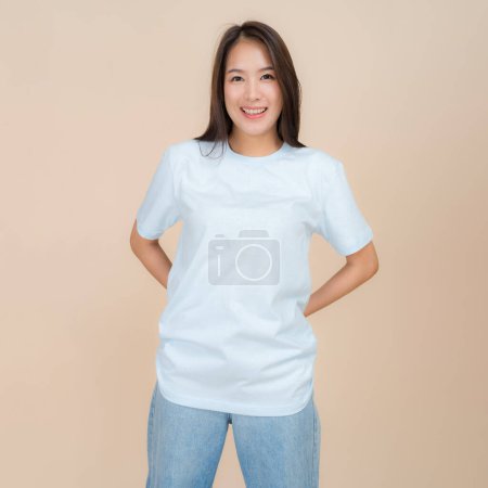 Young asian woman stands confidently against a neutral beige background, wearing a plain blue t-shirt paired with classic blue jeans, representing a simple yet fashionable style