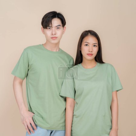 Young asian couple stands side by side wearing matching green t-shirts against a neutral beige background, looking at the camera with a serious expression