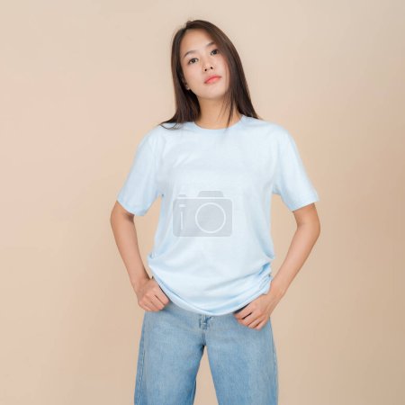 Mujer asiática joven se levanta con confianza contra un fondo beige neutro, usando una camiseta azul liso emparejado con vaqueros azules clásicos, que representan un estilo simple pero de moda