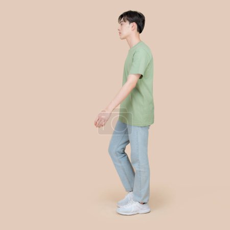 Foto de Vista lateral de un joven asiático de paso medio, caminando casualmente. Lleva una camiseta verde, vaqueros azules y zapatillas blancas contra un fondo beige neutro. - Imagen libre de derechos