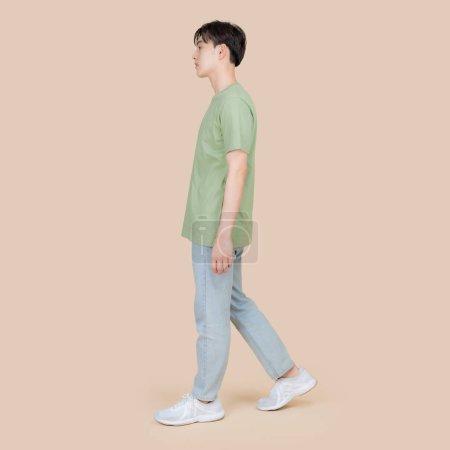 Foto de Vista lateral de un joven asiático de paso medio, caminando casualmente. Lleva una camiseta verde, vaqueros azules y zapatillas blancas contra un fondo beige neutro. - Imagen libre de derechos