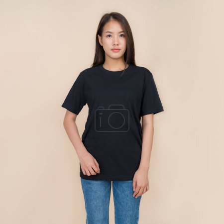 Young asian woman stands confidently against a neutral beige background, wearing a plain black t-shirt paired with classic blue jeans, representing a simple yet fashionable style