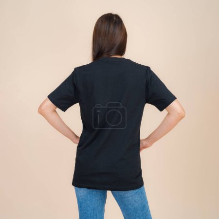 Young asian woman stands confidently against a neutral beige background, wearing a plain black t-shirt paired with classic blue jeans, representing a simple yet fashionable style