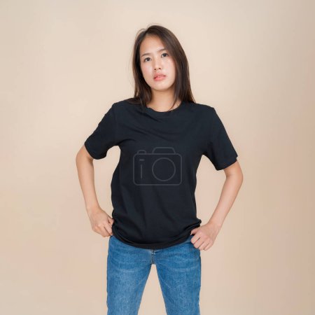 Foto de Mujer asiática joven se levanta con confianza contra un fondo beige neutro, usando una camiseta negra lisa emparejada con vaqueros azules clásicos, que representan un estilo simple pero de moda - Imagen libre de derechos
