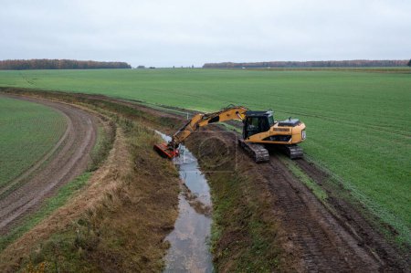 Photographie par drone d'une excavatrice nettoyant un fossé de drainage dans un champ agricole durant la matinée nuageuse d'automne