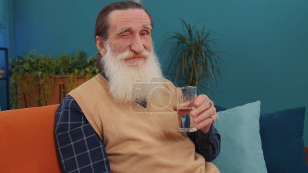 Téléchargez les photos : Portrait d'un homme âgé assoiffé assis à l'intérieur tenant un verre d'aqua naturel faire siroter de l'eau calme empêchant la déshydratation. Grand-père avec de bonnes habitudes de vie, minceur saine, perte de poids - en image libre de droit