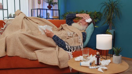 Téléchargez les photos : Vieux grand-père malade âgé souffrant de rhume ou d'allergie couché sur le canapé à la maison. Grand-père malade âgé avec une serviette sur la tête souffle éternuements lingettes morve dans la serviette. Quarantaine de coronavirus, pandémie - en image libre de droit