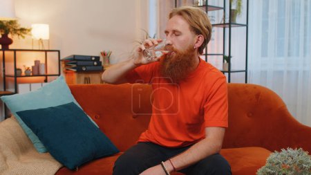 Photo for Thirsty man holding glass of natural aqua make sips drinking still water preventing dehydration. Young guy sitting at home orange couch with good life habits, healthy slimming, weight loss. Lifestyle - Royalty Free Image