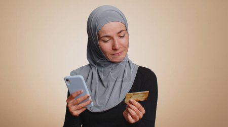 Foto de Mujer musulmana sincera en el cliente hijab utilizando tarjeta de crédito y teléfono inteligente durante la transferencia de dinero, compras en línea de compras, pago. Finanzas e Internet. Chica joven aislada sobre fondo beige - Imagen libre de derechos