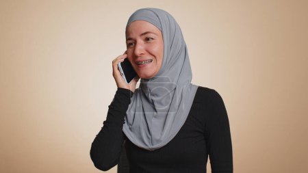 Foto de Feliz joven musulmana mujer en hijab haciendo llamada usando el teléfono celular, hablando agradable conversación con un buen amigo o familia. Arabian chica femenina hablando en el teléfono inteligente en fondo beige estudio - Imagen libre de derechos