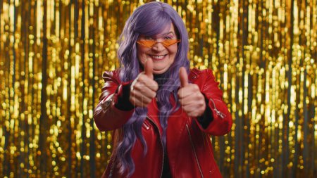 Photo for Like. Stylish elderly woman with purple coiffure raises thumbs up agrees with something or gives positive reply recommends advertisement likes good. Senior grandmother on shiny disco party background - Royalty Free Image