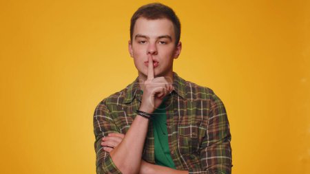 Photo for Shh be quiet please. Portrait of teen adult man 20 years old presses index finger to lips makes silence gesture sign do not tells secret. Young adult handsome boy posing on yellow studio background - Royalty Free Image