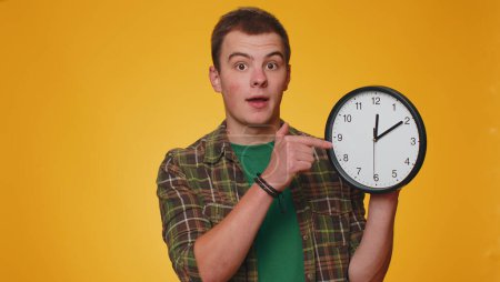 Photo for It is your time. Portrait of teen young man in shirt showing time on clock watch, ok, thumb up, approve, pointing finger at camera. Adult boy indoors studio shot isolated alone on yellow background - Royalty Free Image