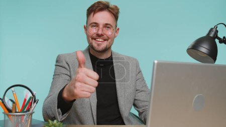 Photo for Like. Happy bearded business man sitting at office desk raises thumbs up agrees with something or gives positive reply recommends advertisement likes good. Young guy boy on blue studio background - Royalty Free Image