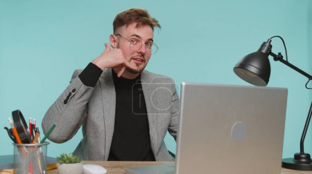 Photo for Call me, here is contact number. Bearded business man sitting at office desk looking at camera doing phone gesture like advertising says hey you call me back. Young guy boy on blue studio background - Royalty Free Image
