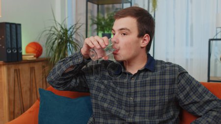 Photo for Portrait of thirsty teenager man sitting indoors holding glass of natural aqua make sips drinking still water preventing dehydration. Guy with good life habits, healthy slimming, weight loss concept - Royalty Free Image