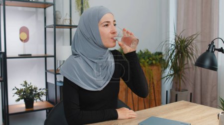 Téléchargez les photos : Une jeune femme musulmane assoiffée en hijab tenant un verre d'aqua naturel fait boire de l'eau calme empêchant la déshydratation. Fille au bureau à domicile avec de bonnes habitudes de vie, un concept sain de perte de poids amincissant - en image libre de droit