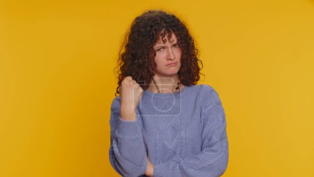 Téléchargez les photos : Aggressive angry curly haired woman trying to fight at camera, shaking fist, boxing with expression, punishment, disappointment, rage, quarrel. Young teen girl isolated on yellow studio background - en image libre de droit