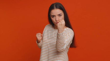 Téléchargez les photos : Angry aggressive pretty woman trying to fight at camera, shaking fist, boxing with expression, punishment, disappointment, rage, quarrel. Young adult girl isolated alone on red studio wall background - en image libre de droit