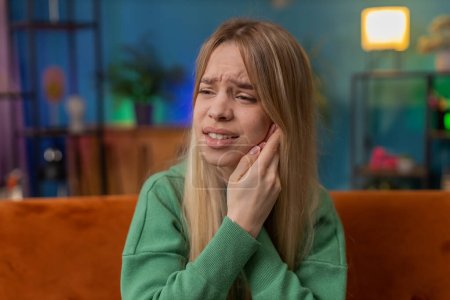 Photo for Dental problems. Young woman touching cheek, closing eyes with expression of terrible suffer from painful toothache, sensitive teeth, cavities. Teen girl at home apartment living room on orange sofa - Royalty Free Image