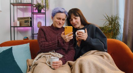 Foto de Two young and cheerful lesbian or friends hold their smartphones while shopping on e-commerce apps at home living room. Girls enjoy having fun browsing different social media applications together - Imagen libre de derechos