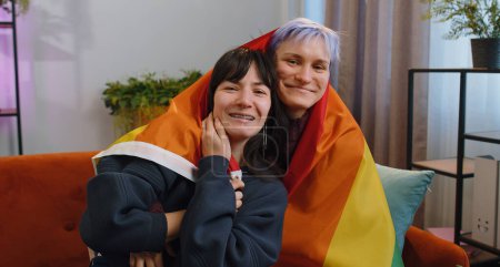 Photo for Homosexual lesbian couple sitting at home room couch next to each other, holding LGBT flag. Smiling girls friends. Pregnant woman from IVF. Lesbian family. Concept of happiness freedom love same-sex - Royalty Free Image