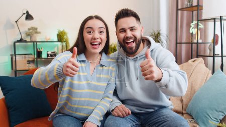 Like. Happy family married couple man woman looking approvingly at camera showing thumbs up, like sign positive something good, positive feedback at home. Husband, wife together on sofa in living room
