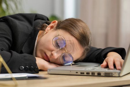 Foto de Aburrido somnoliento mujer de negocios caucásica en gafas de trabajo en el ordenador portátil apoyado en la mano quedarse dormido en la oficina. Chica adicta al trabajo agotada y cansada. Empleo, sin trabajo - Imagen libre de derechos