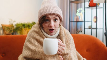 Sick woman wear hat wrapped in plaid sit alone shivering from cold on couch drinking hot tea in unheated apartment without heating due debt. Unhealthy pretty girl feeling discomfort try to warming up