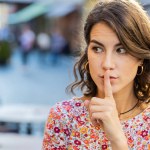 Shh be quiet please. Caucasian young woman presses index finger to lips makes silence hush gesture sign do not tells gossip secret rumors outdoors. Girl standing in urban city street. Town lifestyles