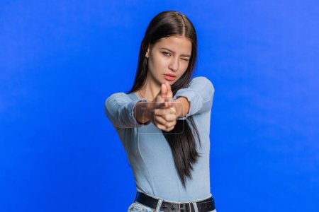 Téléchargez les photos : Une jeune femme caucasienne pointant autour avec un geste de pistolet à doigt, ayant l'air confiante, faisant des choix en tuant avec un pistolet à main sur la cible. Fille isolée sur fond de mur bleu. Publicité - en image libre de droit