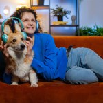 Happy young woman listening to music through wireless headphones lying on couch with corgi dog. Cheerful female girl with curly hair dancing on music in living room at home. Enjoying dance music
