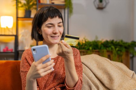 Photo for Portrait of happy woman make payments online purchase shopping internet store. Smiling Caucasian girl holds banking credit card involved in verification process in mobile application on sofa at home - Royalty Free Image