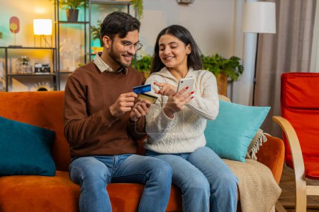 Photo for Happy young couple together doing online shopping by entering credit card number on smartphone while sitting on sofa at home. Smiling family are satisfied with a good promotion on goods in apartment - Royalty Free Image