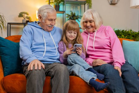Photo for Cute Caucasian girl teaching grandfather and grandmother using smartphone on sofa in living room. Grandparents watching granddaughter play games on mobile phone. Happy family at home during weekend. - Royalty Free Image