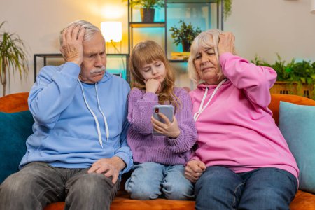 Photo for Upset grandfather, grandmother and granddaughter playing game on smartphone and losing at home. Sad Caucasian girl with grandparents received bad news, fortune loss, fail education study test on couch - Royalty Free Image