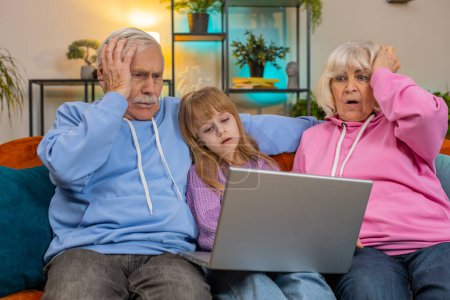 Grand-père aîné bouleversé grand-mère et petite-fille jouer à un jeu sur ordinateur portable et perdre à la maison. Triste fille caucasienne avec des grands-parents a reçu de mauvaises nouvelles perte de fortune échouer test d'étude de l'éducation sur canapé