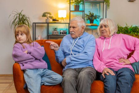 Photo for Irritated grandfather and grandmother scolding granddaughter for bad behavior and disobedience on couch raising voice, scream at little grandchild. Difficulties of upbringing, misbehaved child concept - Royalty Free Image